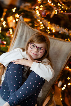 Smiling child, happy little girl relaxing in a chair beside a Christmas tree with warm lights, capturing a cozy real life family moment