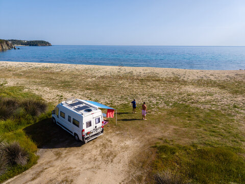 Enjoy a serene getaway at Paralia Azapiko beach in Chalkidiki, Greece. A couple relaxes by their camping van, soaking in the sun and stunning sea views. A perfect holiday escape.