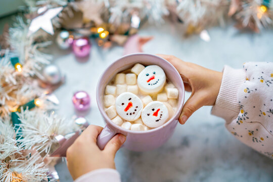 Unrecognizable little child girl holding cup with hot chocolate with marshmallows as snowman. Kid sitting near Christmas decorated window with lights.