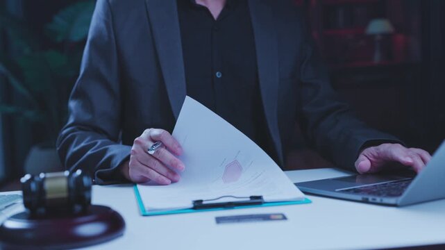 Focused attorney reviewing paper contract documents beside wooden judge gavel while operating portable computer at office desk during evening legal case preparation.