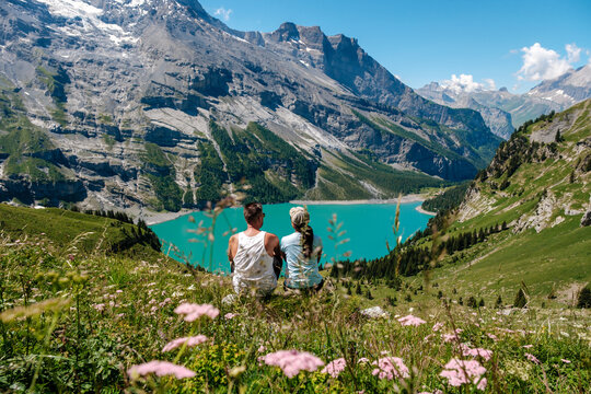 Enjoy the stunning scenery of Oeschinensee in Kandersteg, Switzerland, where two travelers admire the sparkling turquoise lake surrounded by majestic mountains and vibrant alpine flowers.