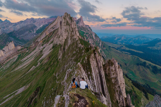 Visitors enjoy breathtaking views from the Ebenalp Schafler ridge in Appenzell, Switzerland. Majestic peaks surround them as the sun sets, casting a warm glow over this beautiful landscape.