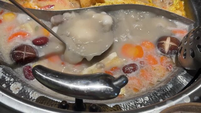 Close up footage of a divided hot pot bubbling with fish fillets, shiitake mushrooms, carrots, red dates and other ingredients as a ladle moves through the broth.