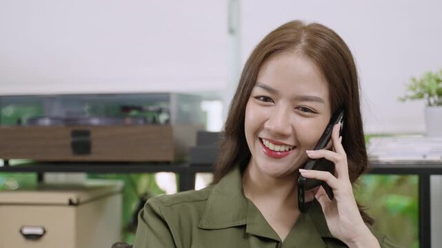 Smiling happy pretty Asian businesswoman sitting working and talking on mobile phone about business with a customer at her desk in office. Young female freelancer using a smartphone in the workplace. 