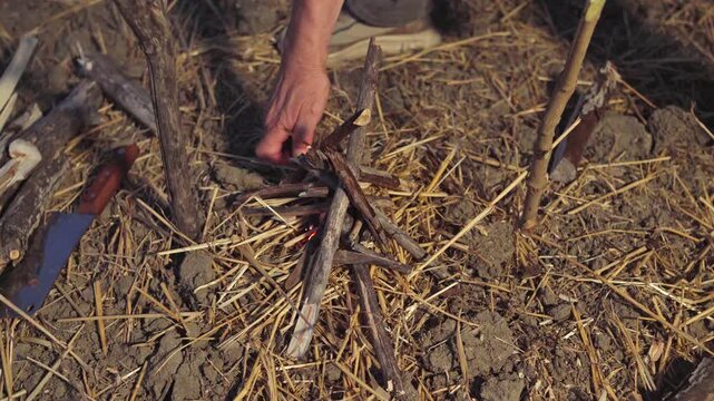 A camper uses a gas lighter to ignite dry kindling sticks and straw in a campfire pit outdoors, close-up shot.