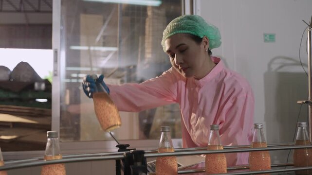 An Asian woman quality control inspector in a uniform of food and drink factory holding a laptop inspecting fruit juice bottles while moving on conveyor belt in beverage production line at industry. 