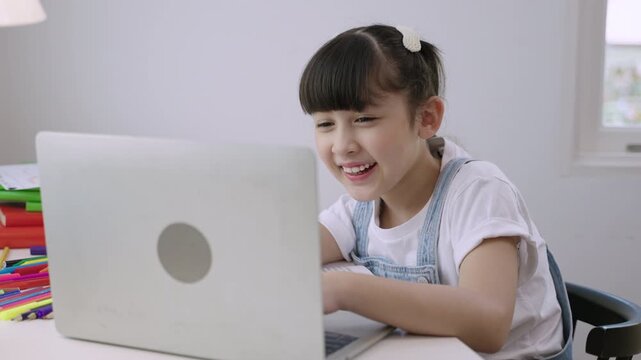 Cute Asian girl student sitting at desk using a laptop computer for studying online learning via video calling with remote teacher in a home school. Homeschooling education and new normal concept. 