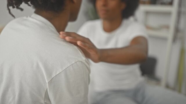 Man places hand on friend's shoulder on a bed in a bedroom, gentle reassuring touch between two african american friends; comfort support.