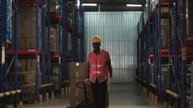 African American male warehouse worker in orange vest uniform wearing yellow safety helmet using hand pallet truck for moving cardboard boxes of cargo from shelves in logistics distribution factory. 