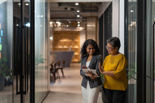 Two cheerful diverse businesswomen of different generations walking in a modern office hallway while discussing a project on a digital tablet.