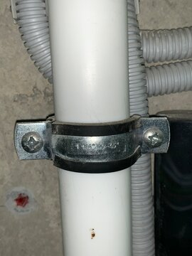 Metal pipe clamp securing white plastic pipe to unfinished concrete wall.
