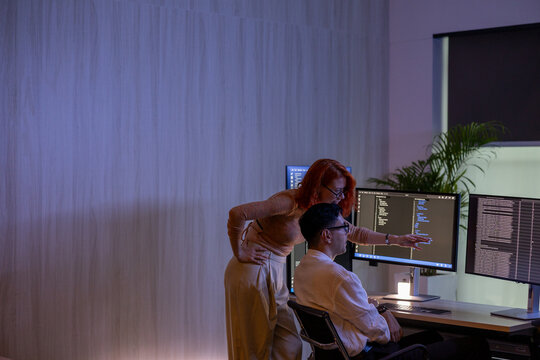 Two focused programmers collaborating on code on multiple computer monitors while working late in a dark office with neon lighting.