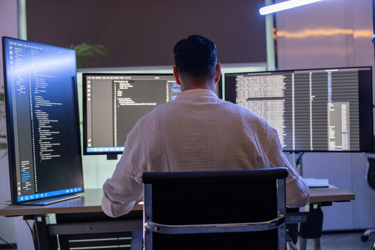 Concentrated young male programmer wearing glasses looking at code on multiple computer monitors in a dark office.