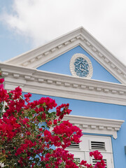 Ornate white pediment and blue gable with decorative roundel above bright pink bougainvillea, classical facade detail with sky backdrop and refined architectural trim © Hernan
