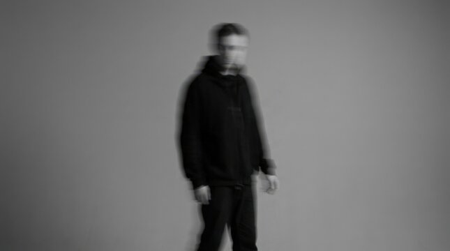 Anonymous person in dark clothing intentionally blurred by motion against a smooth gray background, minimalist conceptual portrait photography, monochrome palette, soft edges, centered composition and