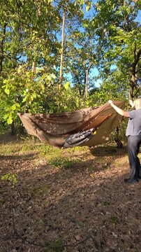 A male camper ties a brown rainfly tarp to a tree in the forest.