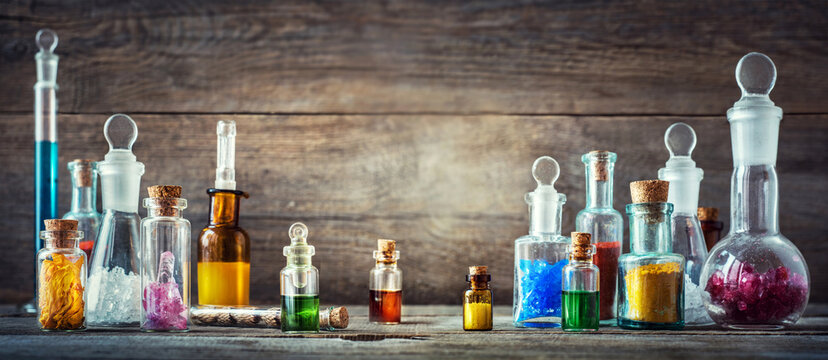 Vintage collection of antique glass apothecary bottles and laboratory vials filled with colorful liquids, crystals, powders, and natural substances on an old wooden table.