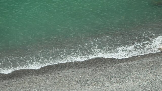 Turquoise ocean waves roll onto a gray gravel shore, creating rhythmic white foam at the water edge. Static high angle overhead shot.
