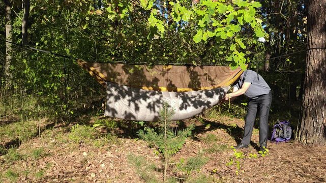 A male camper cinches a hammock underquilt cord in the forest.
