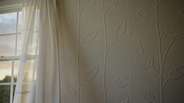 Breeze nudging sheer curtain across sash window at home, revealing embossed leaf-and-vine panel