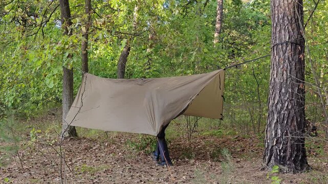 Male camper steps under a pitched tarp shelter in the woods.