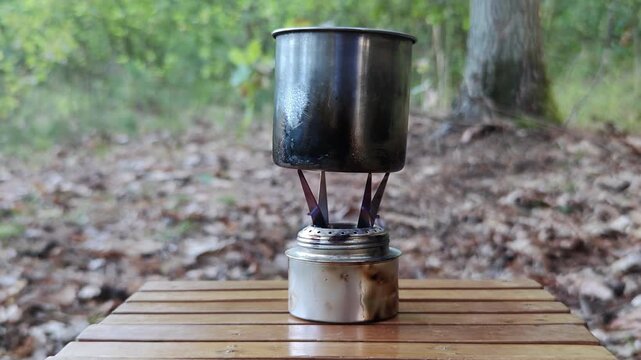 Alcohol stove heats a metal pot on a wooden table in the woods.