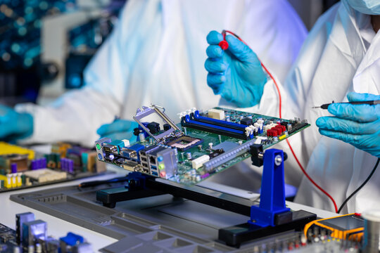 Technicians in white lab coats and blue gloves test a computer motherboard with probes in an electronics manufacturing lab.