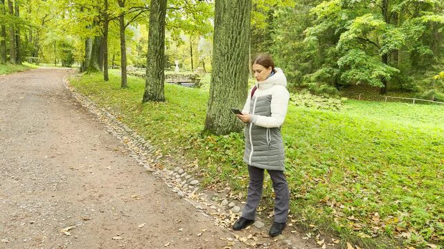 A woman stands looking at her phone along a shaded path in Pavlovsk Park, Saint Petersburg. The autumn trees create a peaceful feeling of quiet calm and relaxation.