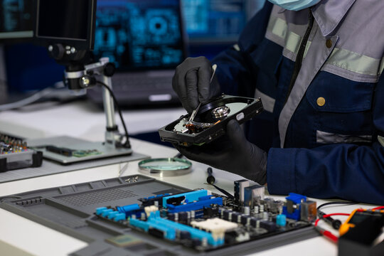 Computer repair technician in gloves examines a hard drive above a motherboard at an electronics workshop with diagnostic screens in the background.
