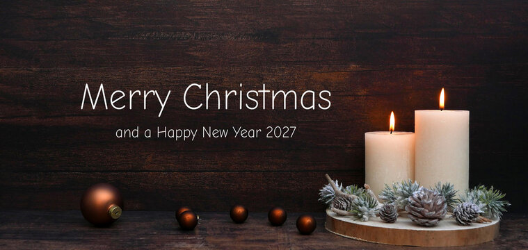 Two burning white candles on a wooden slice with pine cones and Christmas baubles against a dark wooden background, with the text "Merry Christmas and a Happy New Year 2027".