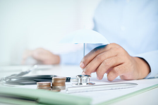 Doctor holding a small umbrella over a stethoscope and coin stacks on a white desk, concept of health insurance coverage, medical care protection, and financial healthcare planning