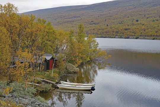Small lake with boats and a hut on the shore, surrounded by autumn trees, mountains in the background, Valasjoen, Dovrefjell-Sunndalsfjella National Park, Norway