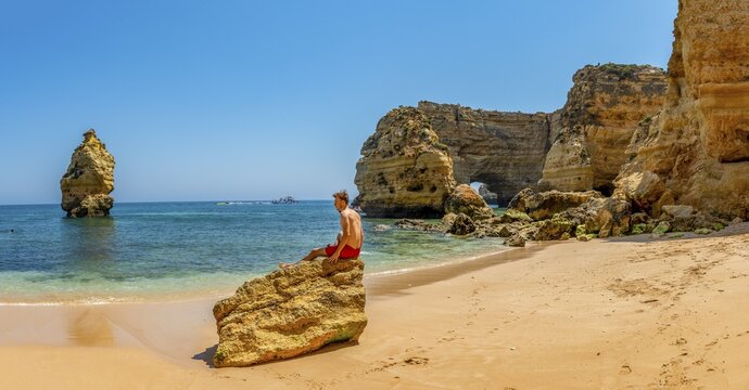 Young man sitting on a rock, Praia da Marinha beach, rocky coast, rock formation in the sea, Algarve, Lagos, Portuga