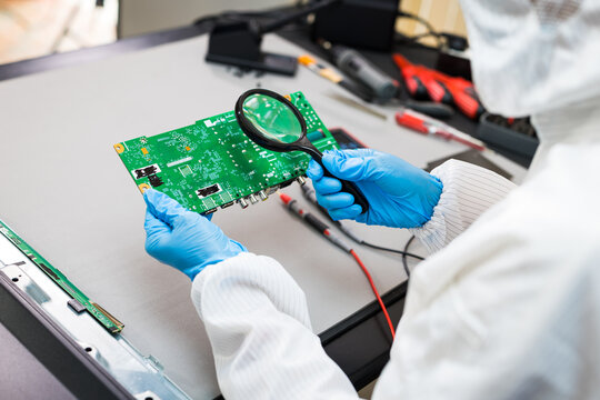Technician in repair clothing and blue gloves inspects a green circuit board with a magnifying glass at an electronics repair workstation.