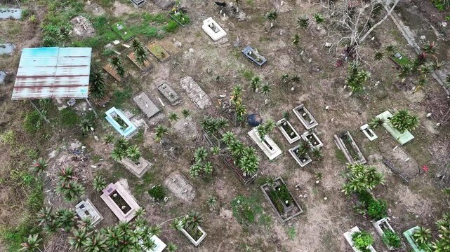 A view from above of a cemetery in Indonesia overgrown with weeds, featuring rectangular graves and a rusty shack.
