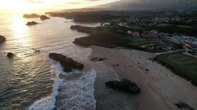 Aerial view of Borizu Beach at sunrise with waves washing over a rocky sea stack, campsites, farmland, and the Sierra del Cuera mountains behind. Llanes, Asturias, Spain