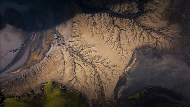Aerial view of intricate brown sand and silt patterns formed by glacial river deltas, with dark water channels and patches of vibrant green moss in Vik, Myrdalshreppur, Iceland.