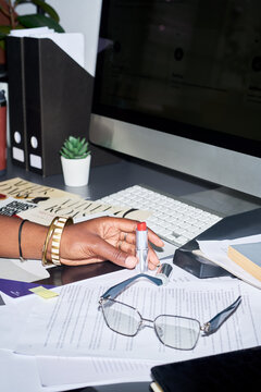 Vertical shot of African American young adult woman holding red lipstick at office desk with computer. Useful for beauty marketing, workplace wellness, and cosmetics branding
