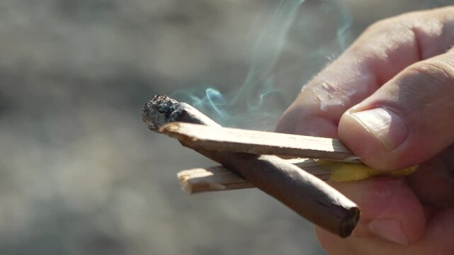Hand holding a smoldering handmade cigar secured with wooden splints outdoors. Close-up macro tracking shot.