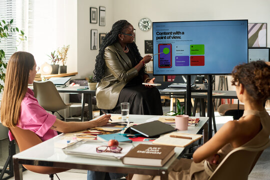 African American middle aged businesswoman presenting design strategy on monitor to Caucasian young adult female colleagues. Image suitable for creative branding and marketing campaign