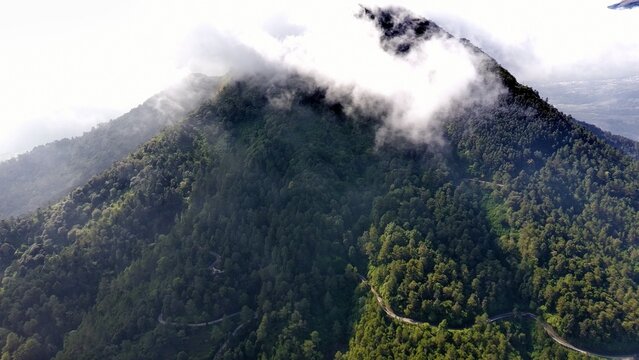 Aerial view of Mount Telomoyo covered in lush green forest with a winding road visible through the dense vegetation and clouds drifting over the peak in Getasan, Central Java, Indonesia.