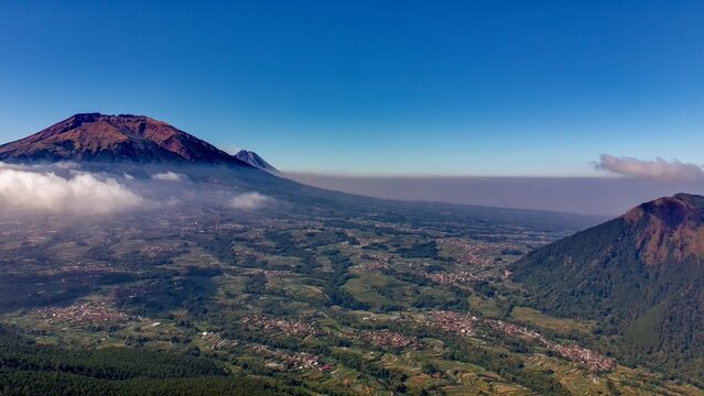 Aerial view of the lush terraced fields and villages nestled at the base of a majestic volcano under a clear blue sky in Ngablak, Central Java, Indonesia.
