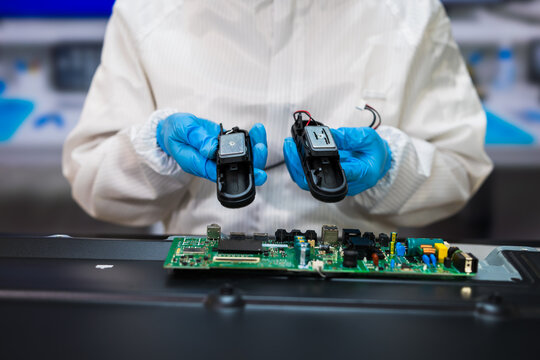 Technician in protective clothing and blue gloves inspects electronic components above a circuit board on an industrial workbench.
