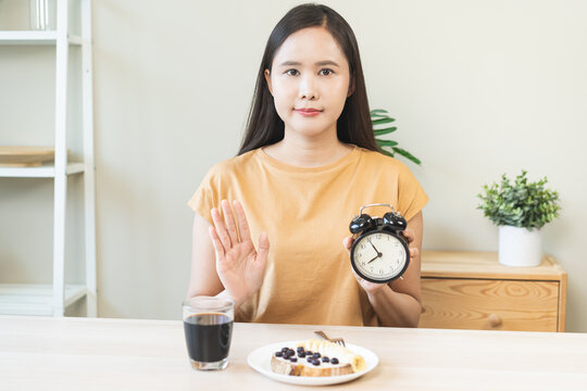 Asian young woman raising hand in stop gesture refusing healthy breakfast food while holding alarm clock, intermittent fasting weight loss regimen and time restricted eating nutrition concept.