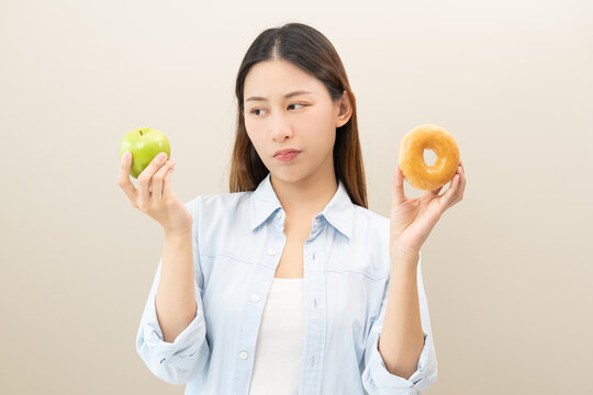 Diet concept, pretty hungry asian young woman, girl confused choose, choice eat food for good healthy, hand holding green apple and sugar sweet donut isolated on background. Temptation of loss weight.