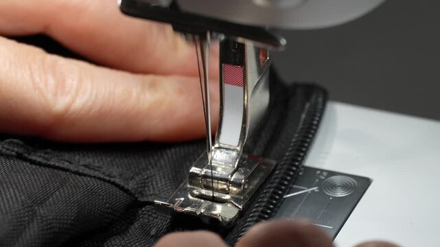 Close-up footage of a woman carefully stitching a black zipper onto fabric using a sewing machine. Detailed view of the needle, thread, zipper and hands during the sewing process.