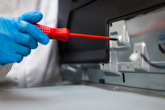 Close-up of a gloved technician using a red screwdriver to repair the metal interior panel of a household appliance in a workshop