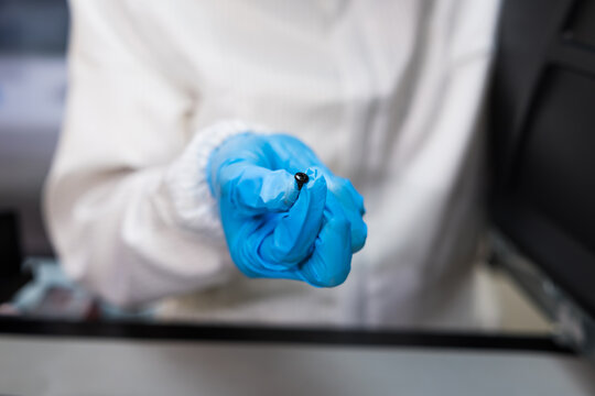 Laboratory worker in a white protective coat and blue glove holds a tiny dark sample over a work surface, suggesting scientific research, testing, safety, and careful handling.