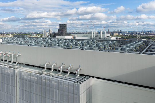 Aerial view of a data center rooftop featuring rows of cooling units and industrial piping with a city skyline in the background, United Kingdom.