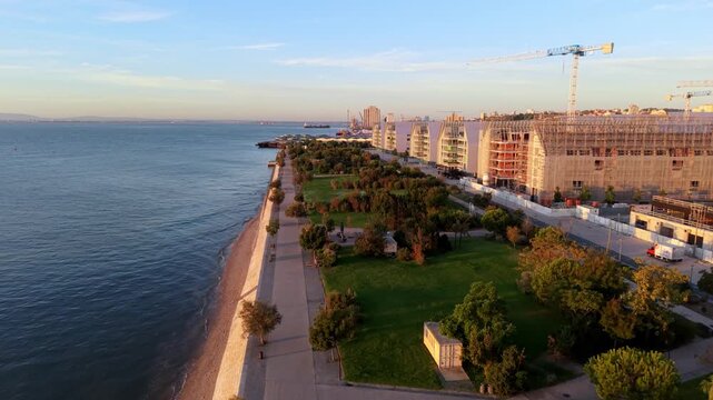 Aerial view of coastal cityscape at golden hour. Modern buildings under construction stretch along seaside promenade. Green parkland separates road from calm ocean waters